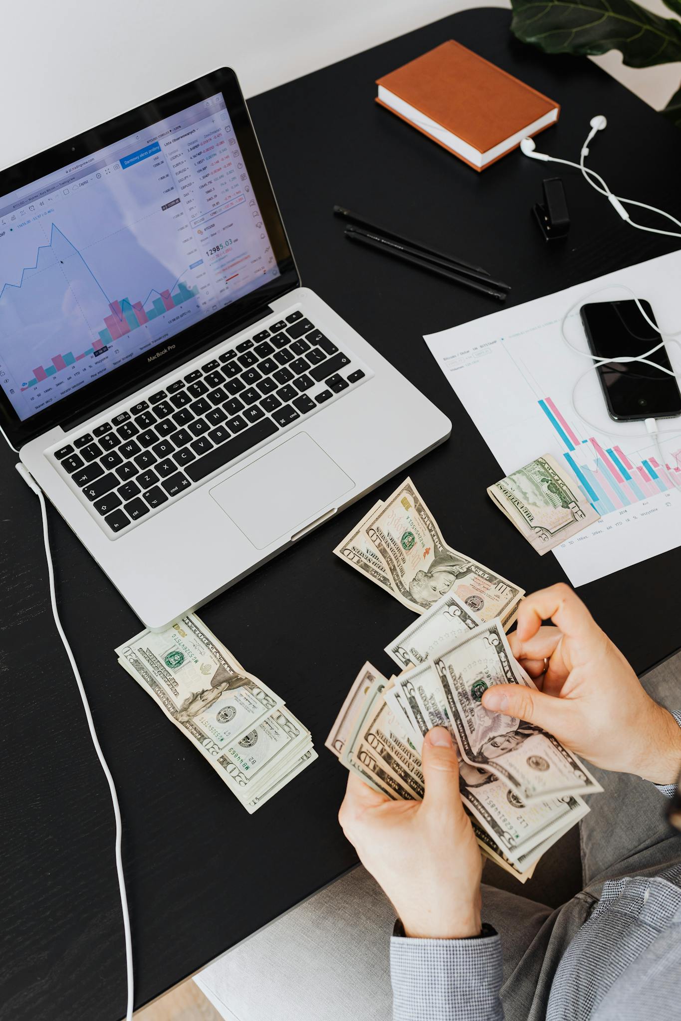 Person counting cash with a laptop analyzing financial trends on a desk.